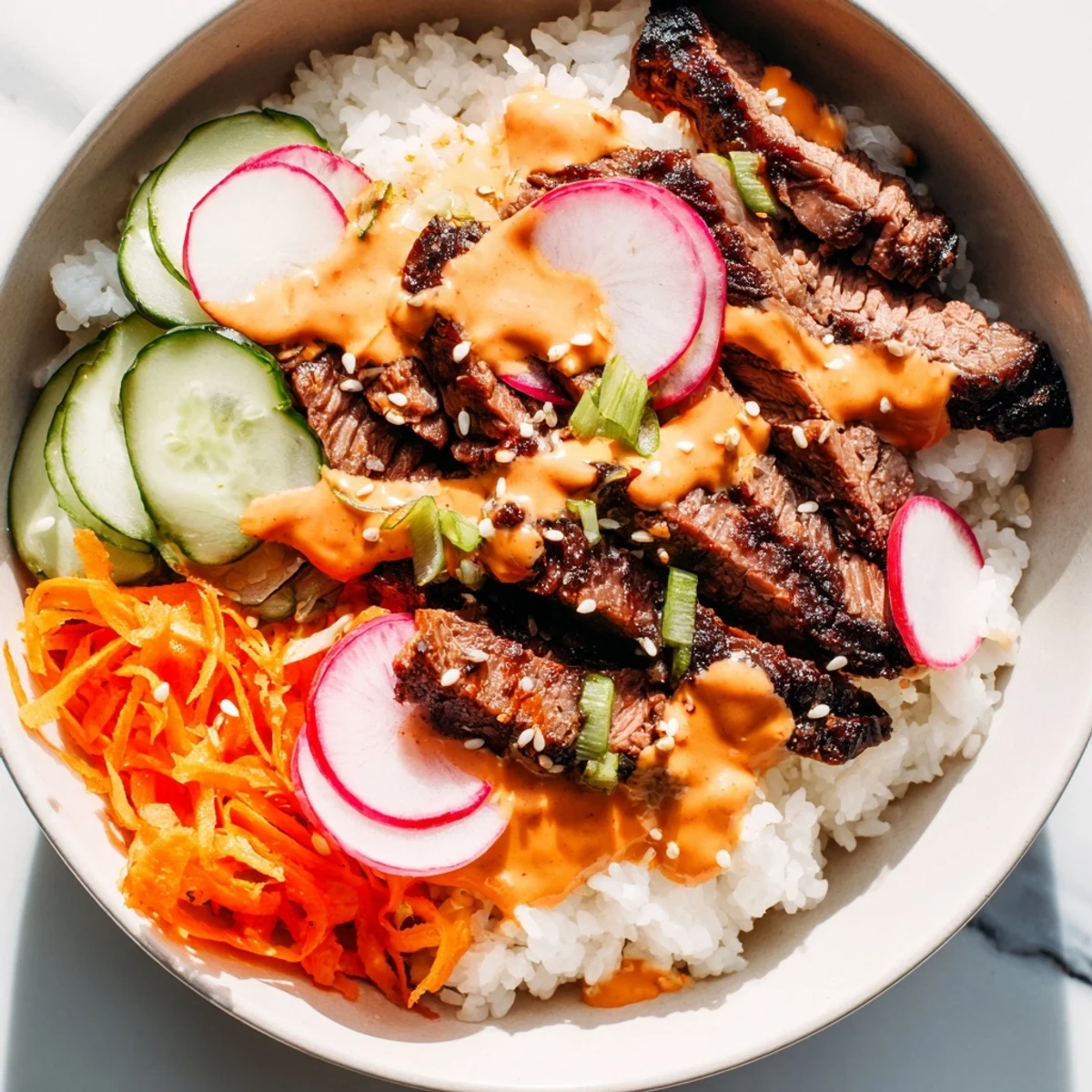 Caramelized marinated flank steak arranged in a bowl with rice cucumbers carrots and creamy spicy sriracha mayonnaise