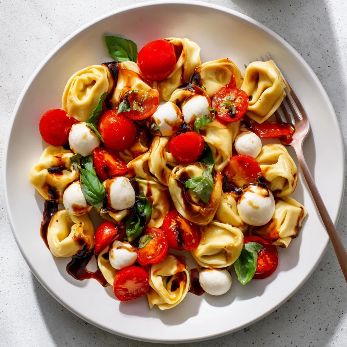 Fresh tortellini Caprese salad drizzled with balsamic glaze alongside halved cherry tomatoes and basil on a rustic wooden table